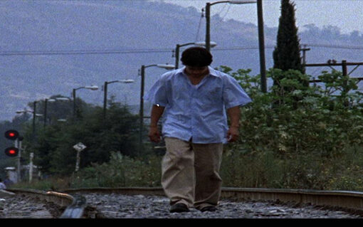 Fotograma de la película Buenas intenciones. un joven con camisa, pantalón y zapatos, anda cabizbajo por las vías de un tren y se ve un tren al fondo.