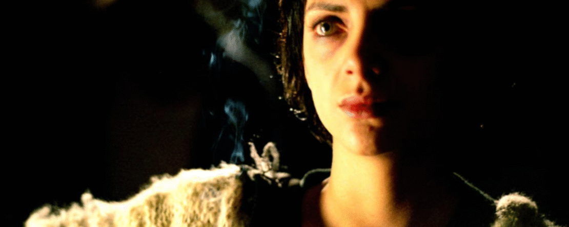 Mujer joven con expresión seria sosteniendo un cigarrillo encendido en un ambiente oscuro.