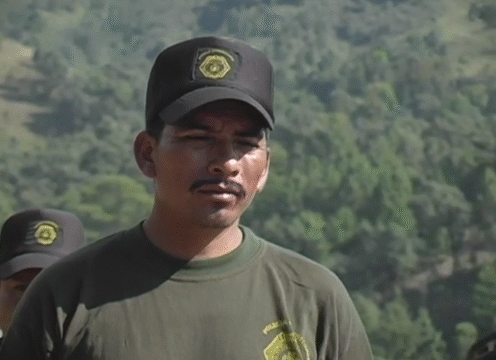 Imagen de uno de los policías, viste la camisa negra y gorra de la policía. Presenta un gesto serio y está mirando al frente. Es un día soleado y al fondo hay un frondoso bosque verde