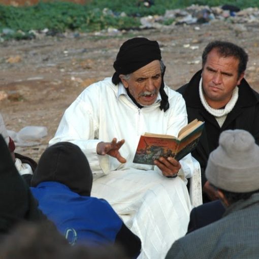 Hombre mayor con túnica blanca y pañuelo negro en la cabeza leyendo un libro a un grupo de hombres sentados al aire libre.
