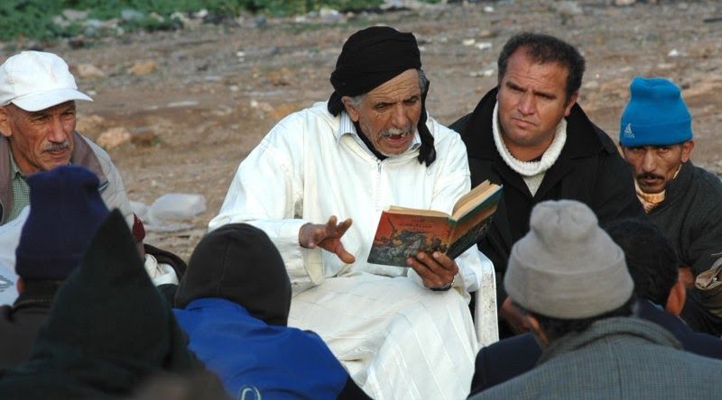 Hombre mayor con túnica blanca y pañuelo negro en la cabeza leyendo un libro a un grupo de hombres sentados al aire libre.