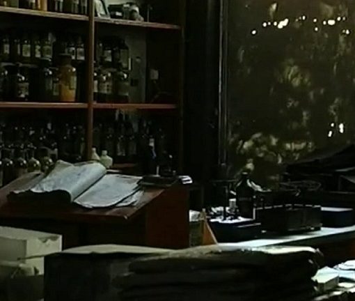 Farmacia antigua con una estantería llena de frascos y un libro abierto. La luz es muy escasa.