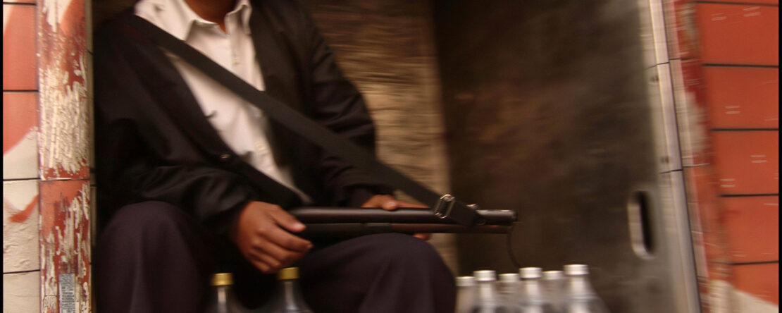 Hombre sentado en la parte trasera de un camión junto a varias botellas de Coca-Cola, sosteniendo un objeto alargado en las manos.