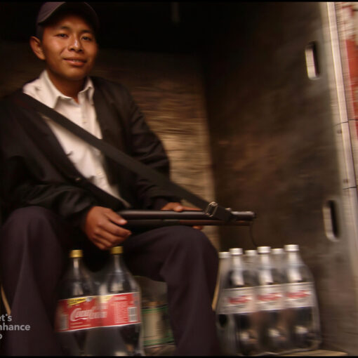 Hombre sentado en la parte trasera de un camión junto a varias botellas de Coca-Cola, sosteniendo un objeto alargado en las manos.