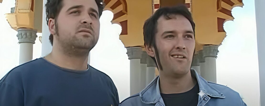 Dos hombres mirando ligeramente a la derecha, con un edificio ornamentado de color claro y arcos al fondo.