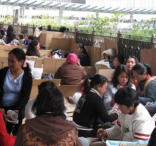 Grupo numeroso de mujeres sentadas en el suelo sobre cartones, compartiendo comida y conversación en un espacio techado al aire libre, rodeado de edificios urbanos.