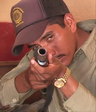 Imagen de voluntario sujetando una pistola y apuntando a cámara, está sentado en un sofa y viste una gorra de la policía una camisa verde y un reloj dorado