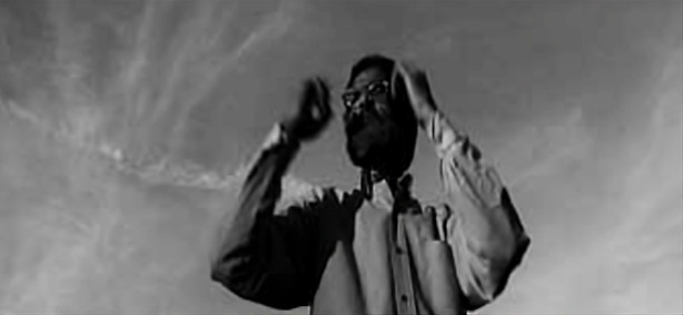 Un hombre barbudo con gafas, visto desde abajo contra un cielo nublado, levanta las manos a la cabeza en un gesto dramático.