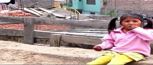 Fotograma de la película el milagro de la pupuna y los aviones. Una niña sentada en el suelo cerca de una cerca, atrás de ella se visualiza el pueblo, chupándose la mano