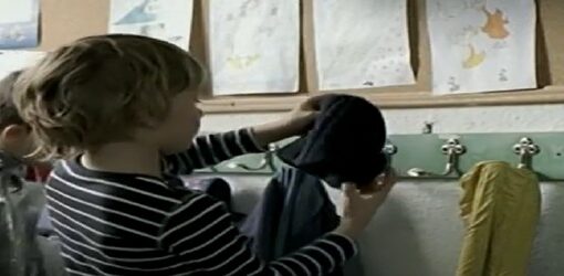 Un niño se pone su gorro frente a un perchero en una pared decorada con dibujos infantiles. La escena parece ocurrir en una escuela o guardería.