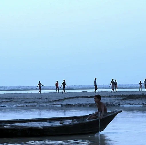 Fotograma en el que aparece niños subidos en unas barcas en un lago. Al fondo, hay más niños corriendo por la arena.