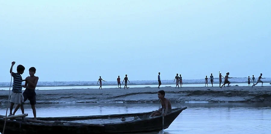Fotograma en el que aparece niños subidos en unas barcas en un lago. Al fondo, hay más niños corriendo por la arena.
