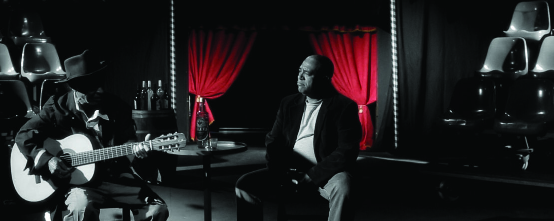 Fragmento del corto - Dos músicos, uno de ellos guitarra en mano, en un salón de actos. Blanco y negro y telón trasero rojo.