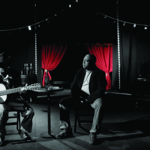 Fragmento del corto - Dos músicos, uno de ellos guitarra en mano, en un salón de actos. Blanco y negro y telón trasero rojo.