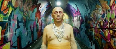 Fotograma de El viaje de Monalisa en el que aparece una persona maquillada en tonos rosa, con un collar muy grande de perlas y sin camiseta. Está en un túnel lleno de grafitis de colores.