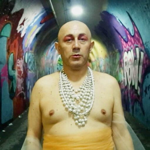 Fotograma de El viaje de Monalisa en el que aparece una persona maquillada en tonos rosa, con un collar muy grande de perlas y sin camiseta. Está en un túnel lleno de grafitis de colores.