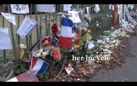 Homenaje callejero con flores, mensajes, velas y banderas francesas junto a una bicicleta decorada, en memoria de una víctima. En la imagen se lee "her bicycle".