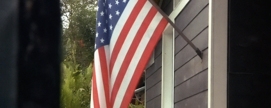 Imagen de una bandera de estados unidos instalada en el porche de una casa. La casa es de madera y está pintada de color negro con una ventana blanca, al fondo hay un bosque frondoso y el cielo aunque claro, está nublado.
