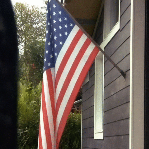 Imagen de una bandera de estados unidos instalada en el porche de una casa. La casa es de madera y está pintada de color negro con una ventana blanca, al fondo hay un bosque frondoso y el cielo aunque claro, está nublado.