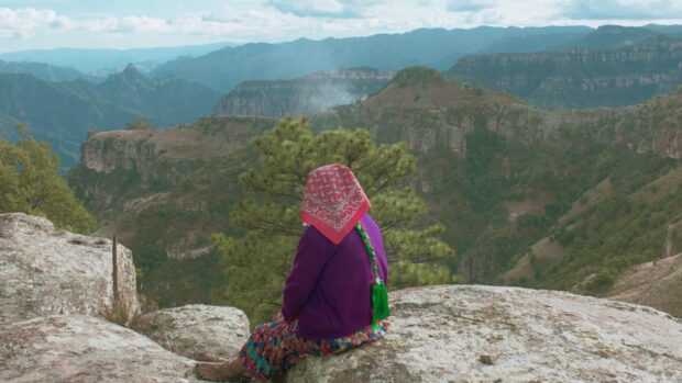 Imagen de una persona de espaldas sentada en una roca en lo alto de un mirador observando un verde y profundo valle. Viste falda colorida, una chaqueta de punto morada y un colorido pañuelo rosa en la cabeza