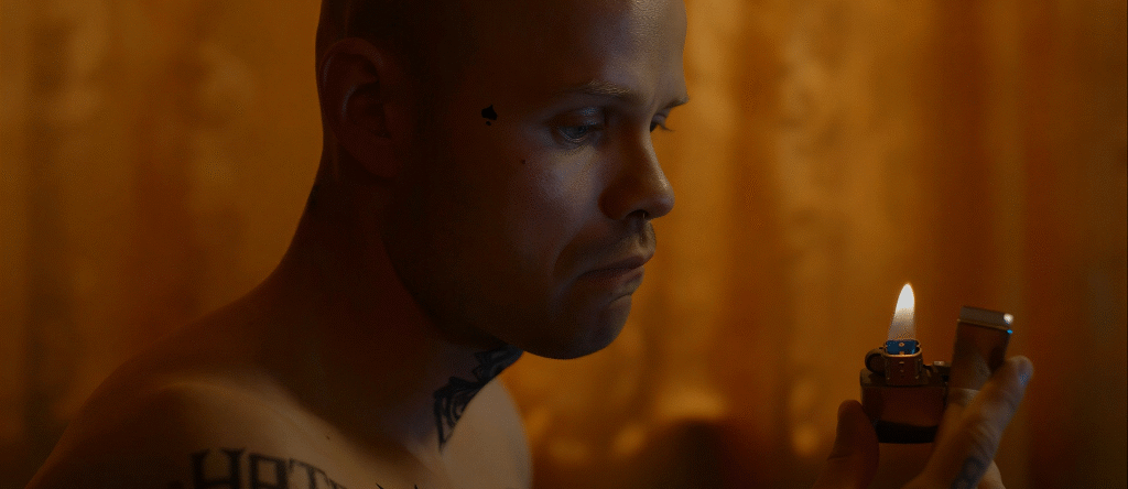 Primer plano del protagonista, esta sin camiseta y se pueden ver sus tatuajes. Está sujetando un mechero que mira fijamente. El fondo desenfocado parece una cortina color naranja dentro de una habitación.