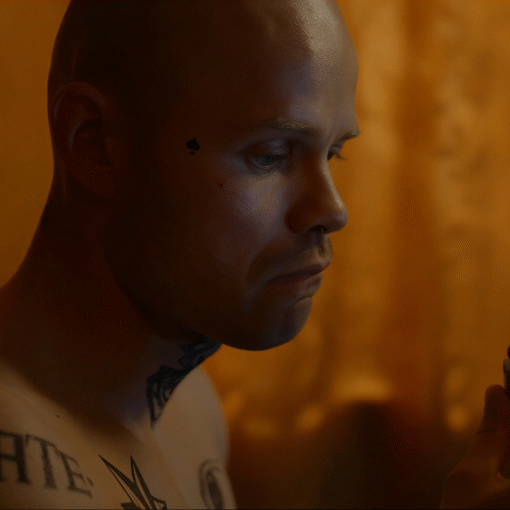 Primer plano del protagonista, esta sin camiseta y se pueden ver sus tatuajes. Está sujetando un mechero que mira fijamente. El fondo desenfocado parece una cortina color naranja dentro de una habitación.