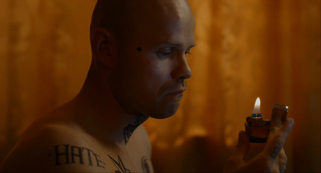 Primer plano del protagonista, esta sin camiseta y se pueden ver sus tatuajes. Está sujetando un mechero que mira fijamente. El fondo desenfocado parece una cortina color naranja dentro de una habitación.