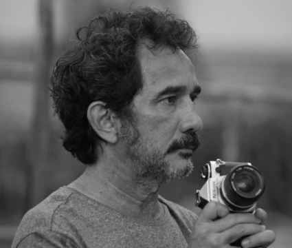 Imagen en banco y negro de un hombre de mediana edad, tiene barba, bigote y el pelo revuelto. Está sujetando con sus manos su cámara de fotos y tiene la mirada fijada en el horizonte. El fondo de la imagen está desenfocado.