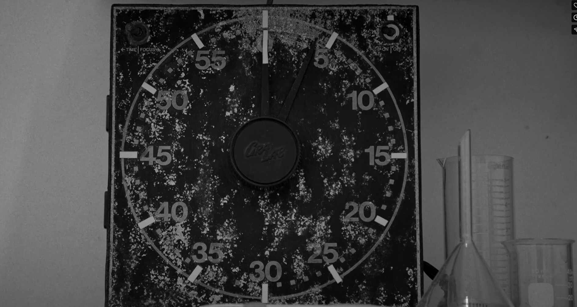 Imagen en banco y negro de un temporizador metálico en muy mal estado toda la platina se ve carcomida por el oxido. A la derecha del temporizar hay una probeta y un vaso de precipitados ambos vacíos.