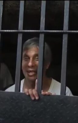 Hombre asomado detrás de unos barrotes metálicos, mirando hacia la cámara con expresión sonriente y sujetándose a la reja con las manos.