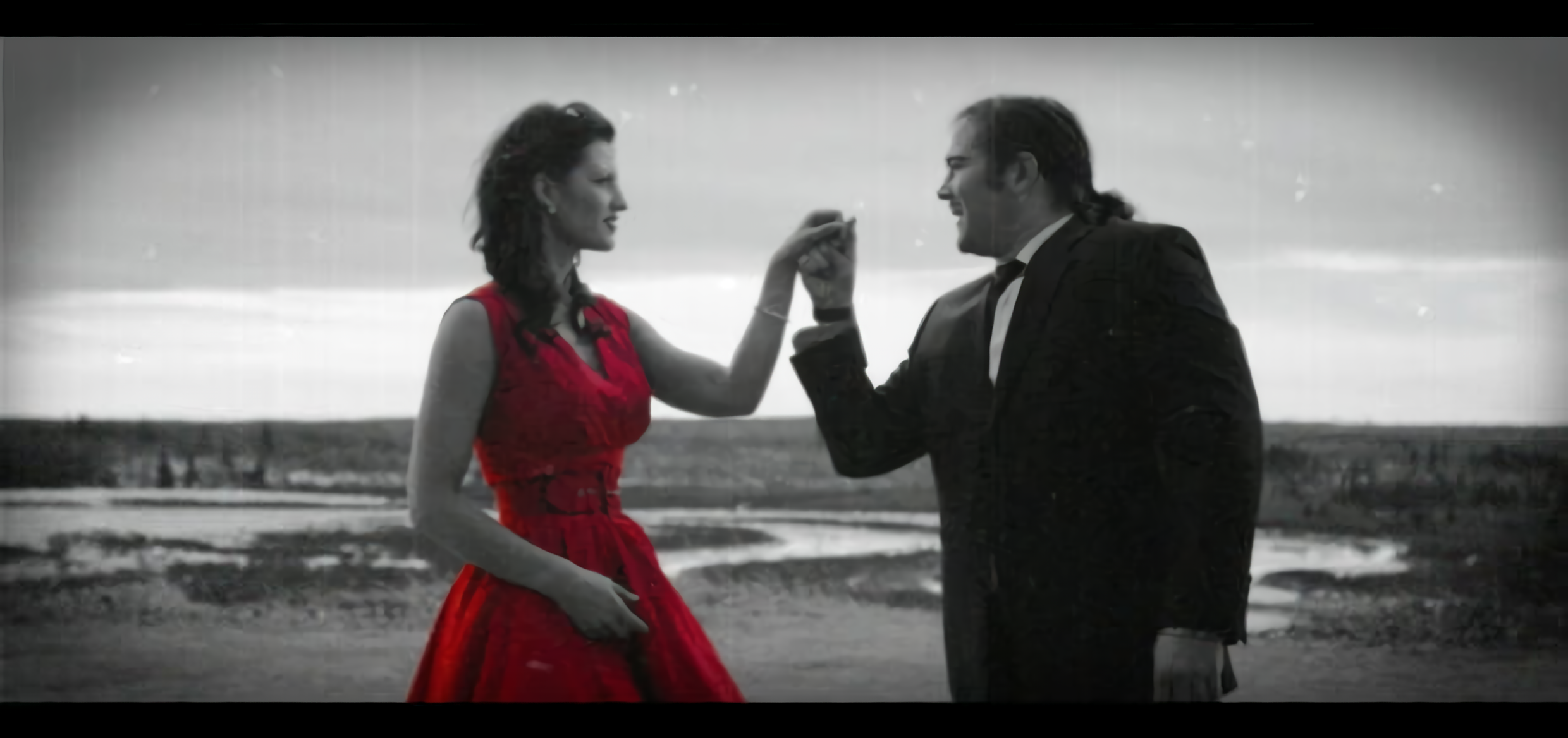 Pareja tomada de la mano en un paisaje abierto, con imagen en blanco y negro y vestido rojo resaltado en color.