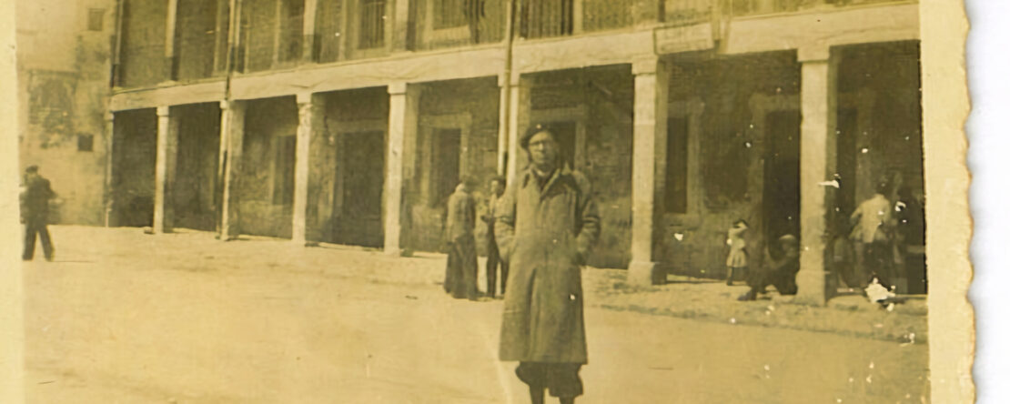 Fotografía antigua en tono sepia de un hombre de pie en una calle frente a un edificio con soportales y balcones.