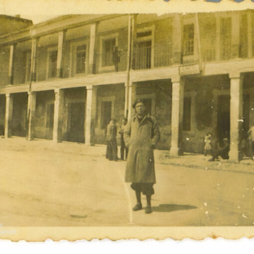 Fotografía antigua en tono sepia de un hombre de pie en una calle frente a un edificio con soportales y balcones.