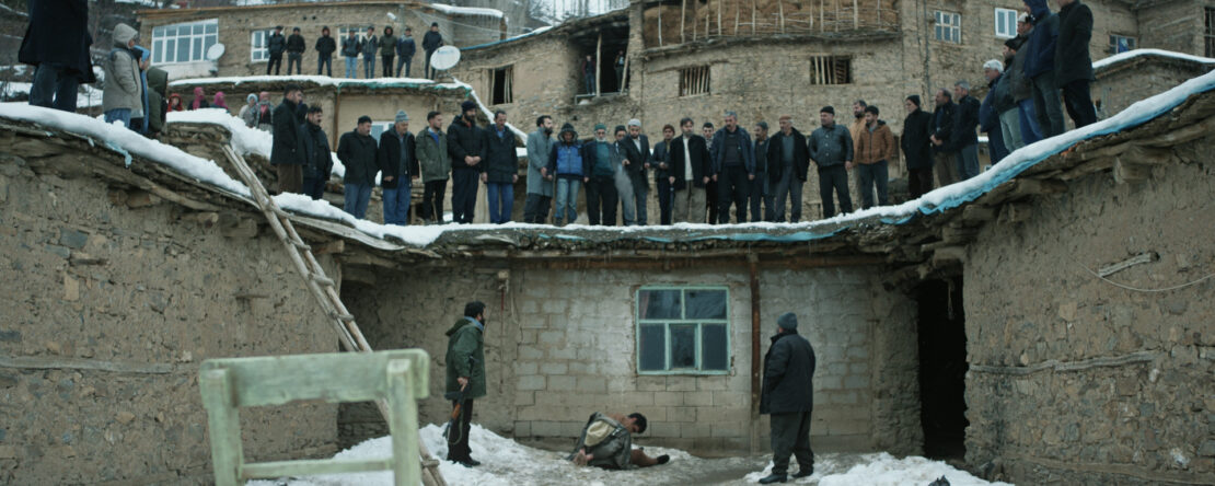 Fotograma de El Destierro. Un hombre está arrodillado en la nieve frente a una casa de adobe, mientras varios aldeanos lo observan desde lo alto de un tejado y el patio. La escena transmite tensión, aislamiento y juicio colectivo en un entorno rural y helado