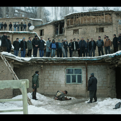 Fotograma de El Destierro. Un hombre está arrodillado en la nieve frente a una casa de adobe, mientras varios aldeanos lo observan desde lo alto de un tejado y el patio. La escena transmite tensión, aislamiento y juicio colectivo en un entorno rural y helado
