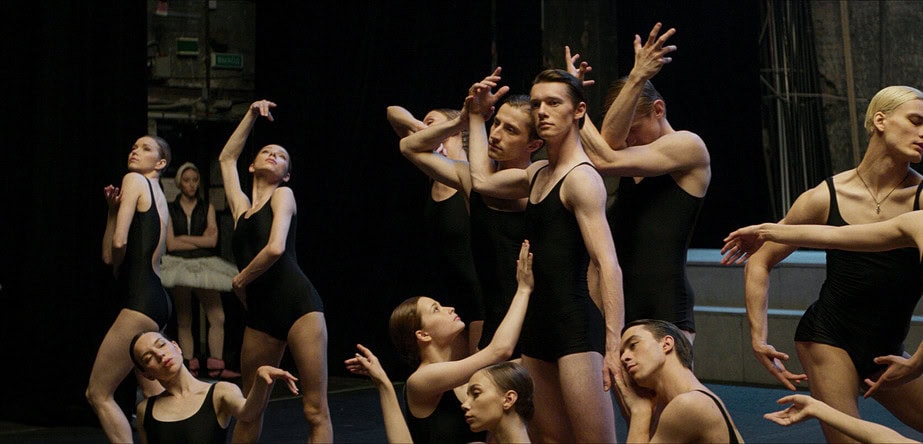 Un grupo de bailarines de ballet vestidos con mallas negras y trajes de práctica, algunos realizando poses con los brazos elevados. Se aprecia un escenario oscuro de fondo.