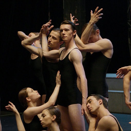 Un grupo de bailarines de ballet vestidos con mallas negras y trajes de práctica, algunos realizando poses con los brazos elevados. Se aprecia un escenario oscuro de fondo.