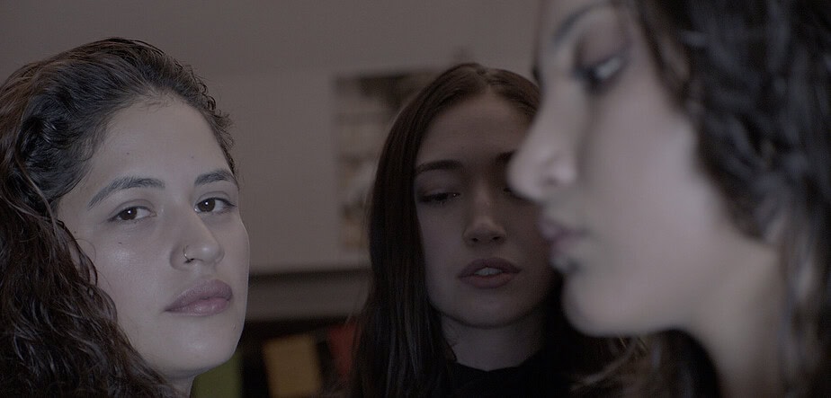 Tres mujeres en un primer plano. Una mira directamente a la cámara mientras las otras dos están de perfil o con la mirada baja.