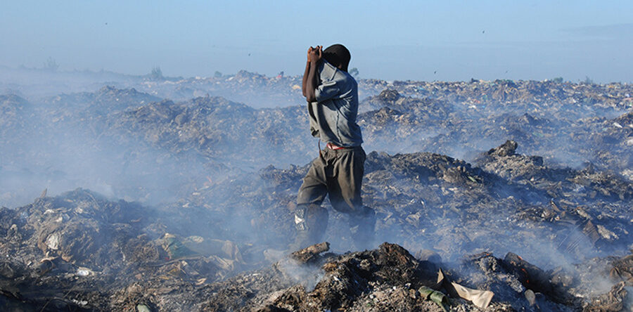 Una persona de espaldas, con los brazos alzados, de pie en un paisaje de vertedero lleno de basura y humo. El cielo es azul y despejado.