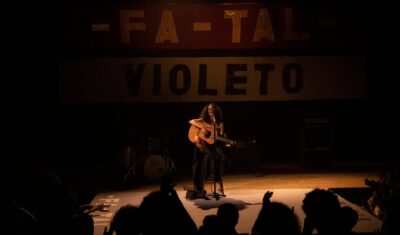 Gal Costa en un escenario oscuro, sentada en un taburete y tocando una guitarra acústica. Al fondo, se ve un telón con las palabras "FA-TAL" y "VIOLETO".