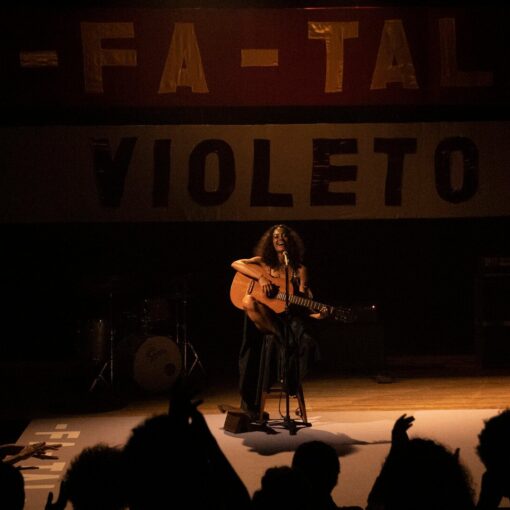 Gal Costa en un escenario oscuro, sentada en un taburete y tocando una guitarra acústica. Al fondo, se ve un telón con las palabras "FA-TAL" y "VIOLETO".