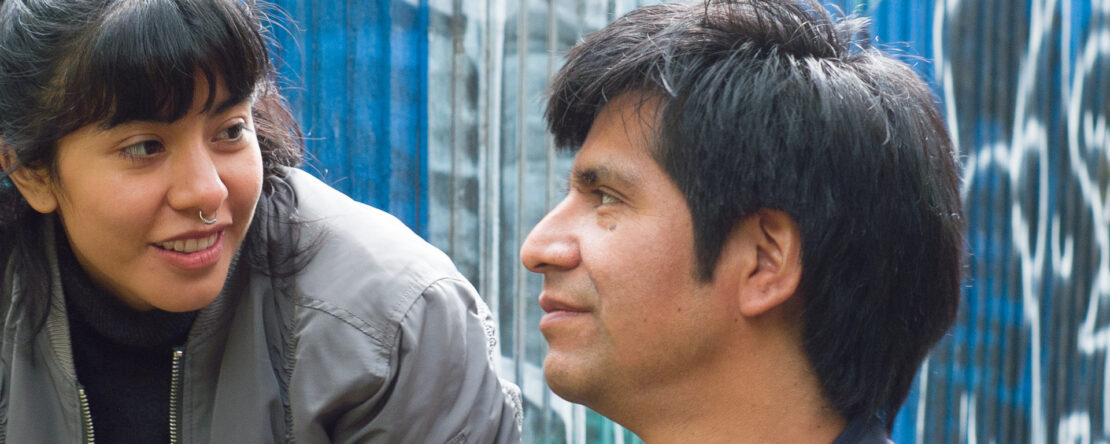 Una joven con piercing y un hombre se observan sonrientes en un entorno urbano con paredes azules. La escena transmite cercanía y humanidad en un mundo hostil.