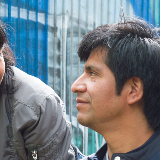 Una joven con piercing y un hombre se observan sonrientes en un entorno urbano con paredes azules. La escena transmite cercanía y humanidad en un mundo hostil.