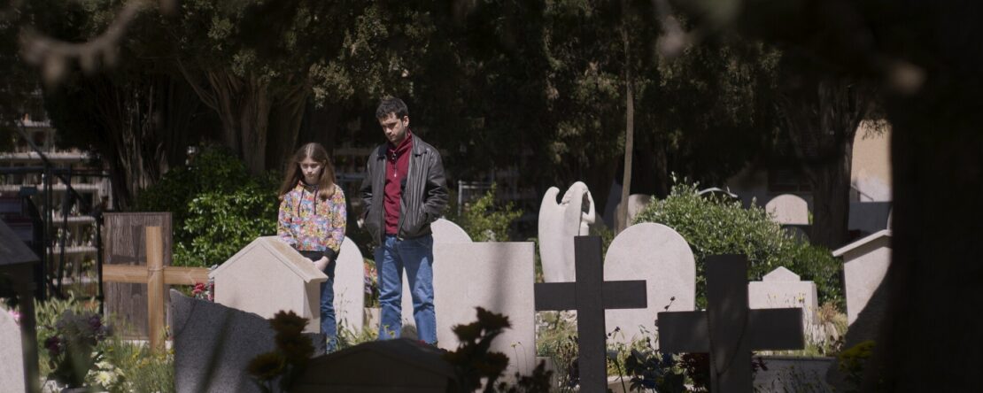 Un hombre y una niña de pie en un cementerio entre lápidas y árboles.