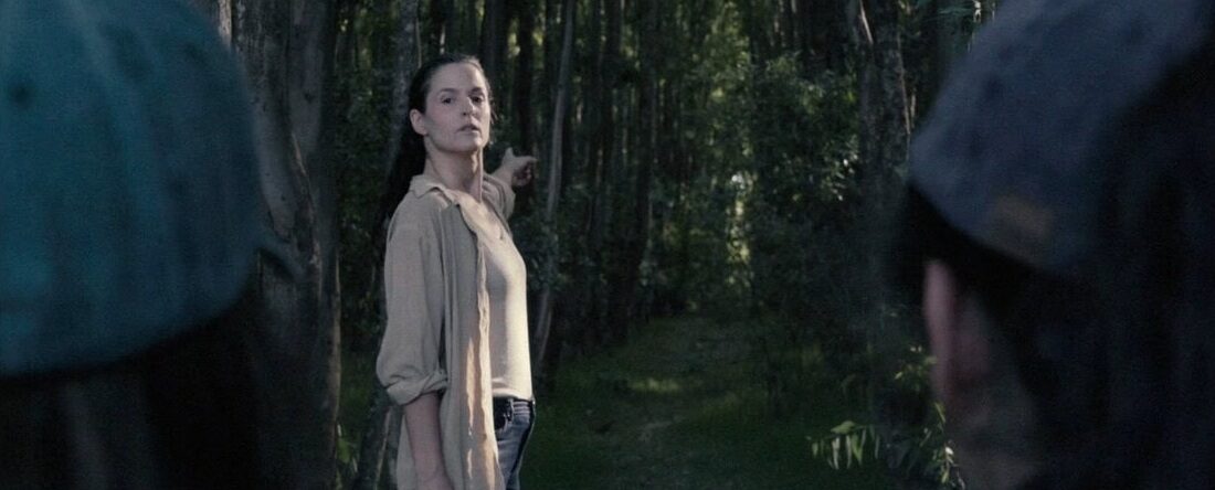 Fotograma del cortometraje No piensen en mí. Una mujer adulta, de pie entre árboles altos, mira hacia atrás mientras señala un sendero. Frente a ella, parcialmente desenfocadas, aparecen dos figuras femeninas. La escena transmite tensión emocional, recuerdo y búsqueda interior en un entorno natural.