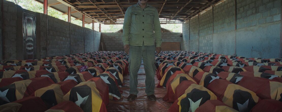 Hombre de pie en un almacén junto a decenas de gallos cubiertos con telas con los colores de la bandera de Timor-Leste. La imagen transmite solemnidad.