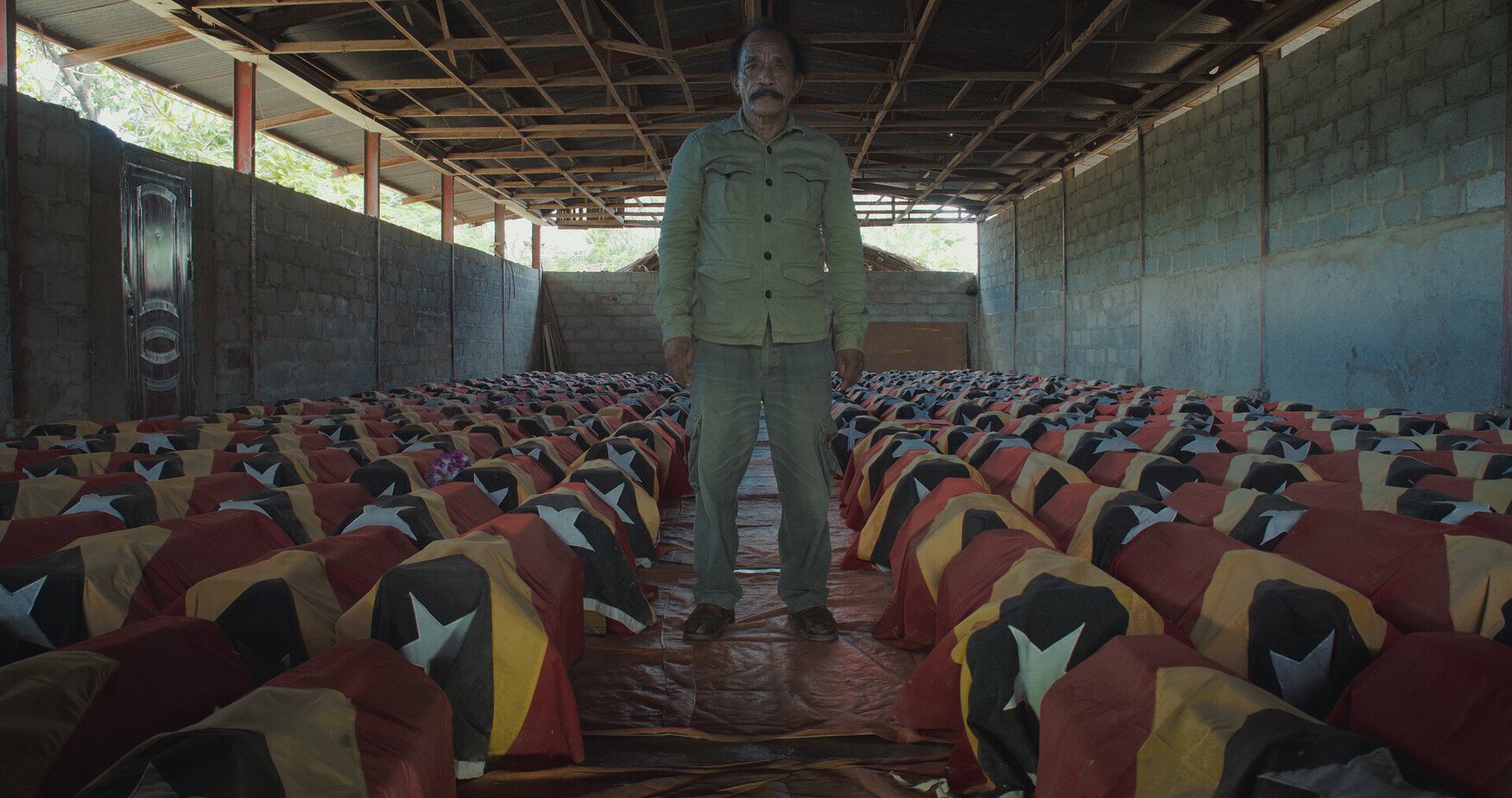Hombre de pie en un almacén junto a decenas de gallos cubiertos con telas con los colores de la bandera de Timor-Leste. La imagen transmite solemnidad.