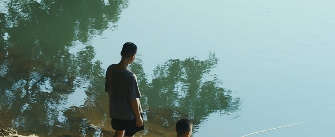 Dos personas jóvenes junto a un cuerpo de agua tranquilo, una de pie y la otra sentada pescando. El reflejo de los árboles se ve en el agua.