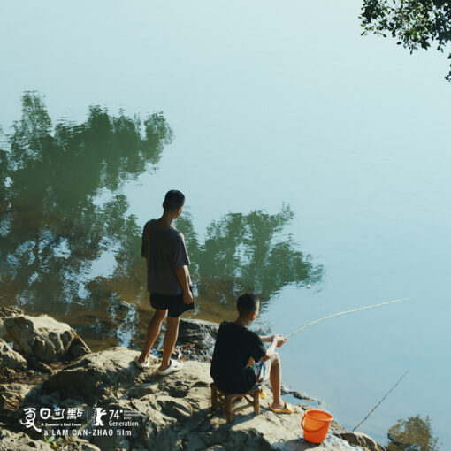 Dos personas jóvenes junto a un cuerpo de agua tranquilo, una de pie y la otra sentada pescando. El reflejo de los árboles se ve en el agua.