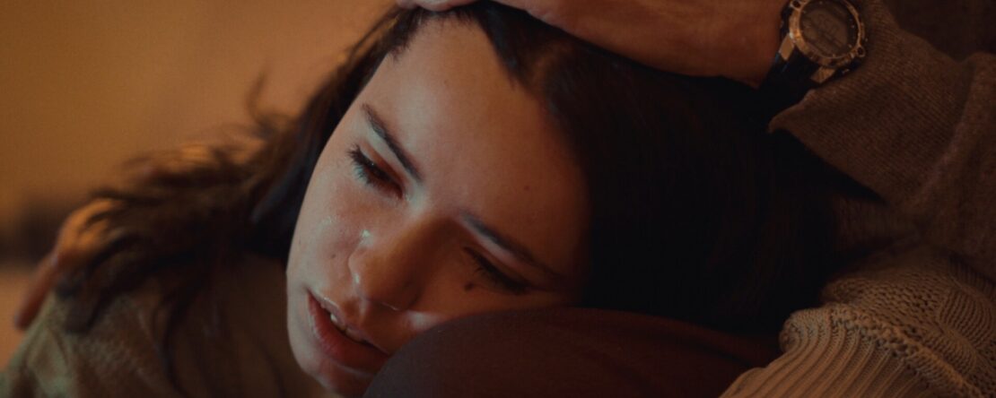 Delia, una joven con el rostro visiblemente emocionado, apoya la cabeza en el regazo de alguien mientras una mano adulta le acaricia con ternura el cabello. La escena está iluminada con luz cálida y transmite un momento íntimo de consuelo y vulnerabilidad emocional.